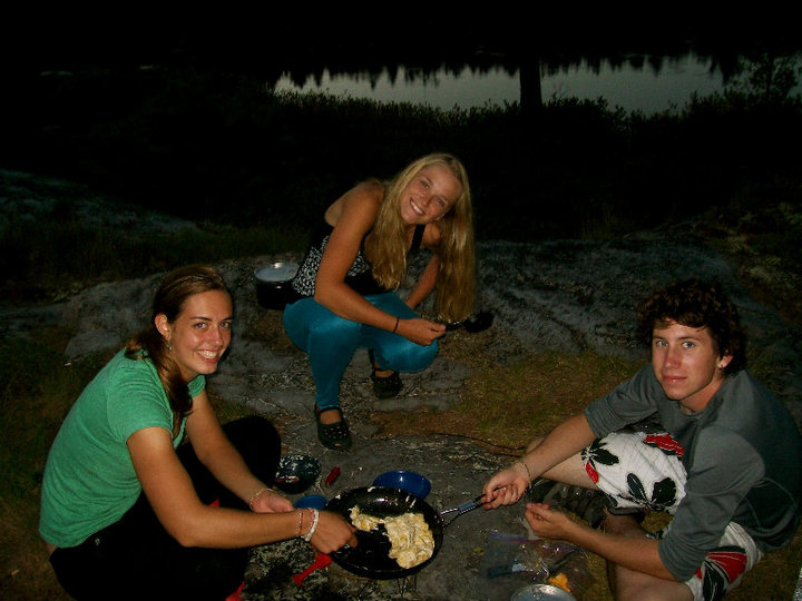 Three individuals prepare a meal over the camp-stoves.