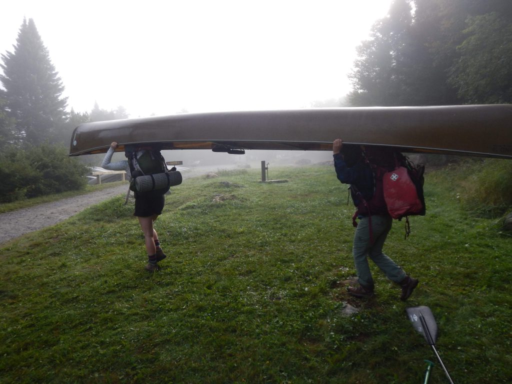 Two individuals portage a canoe with their packs. 