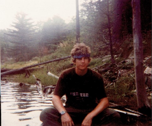 Individual in a bandana sits at the side of a river. 